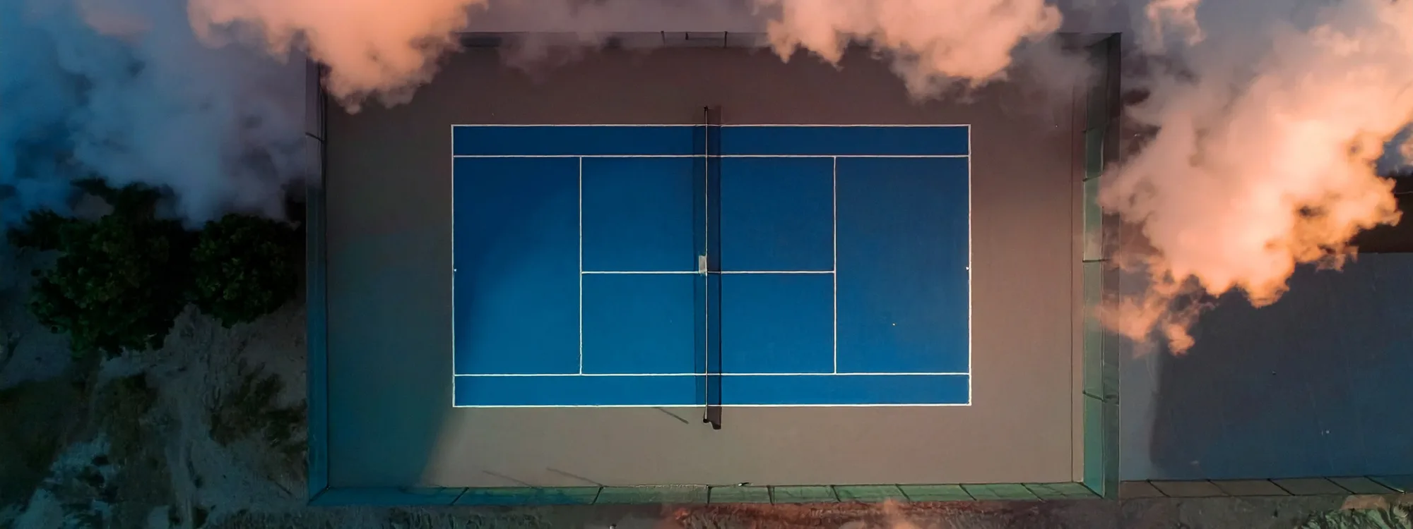 A top-down aerial of a single blue tennis court framed by dramatic clouds at dusk.